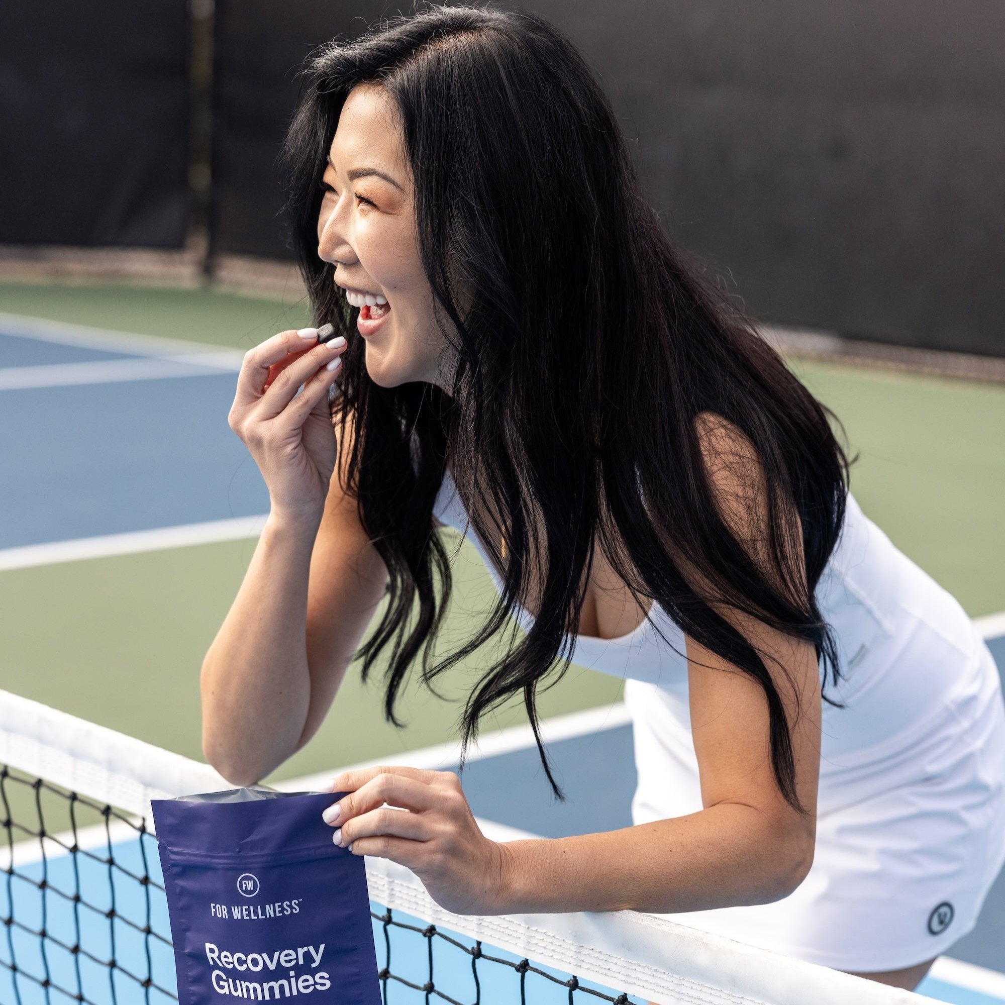 Smiling woman on a tennis court holding recovery gummies by the net.