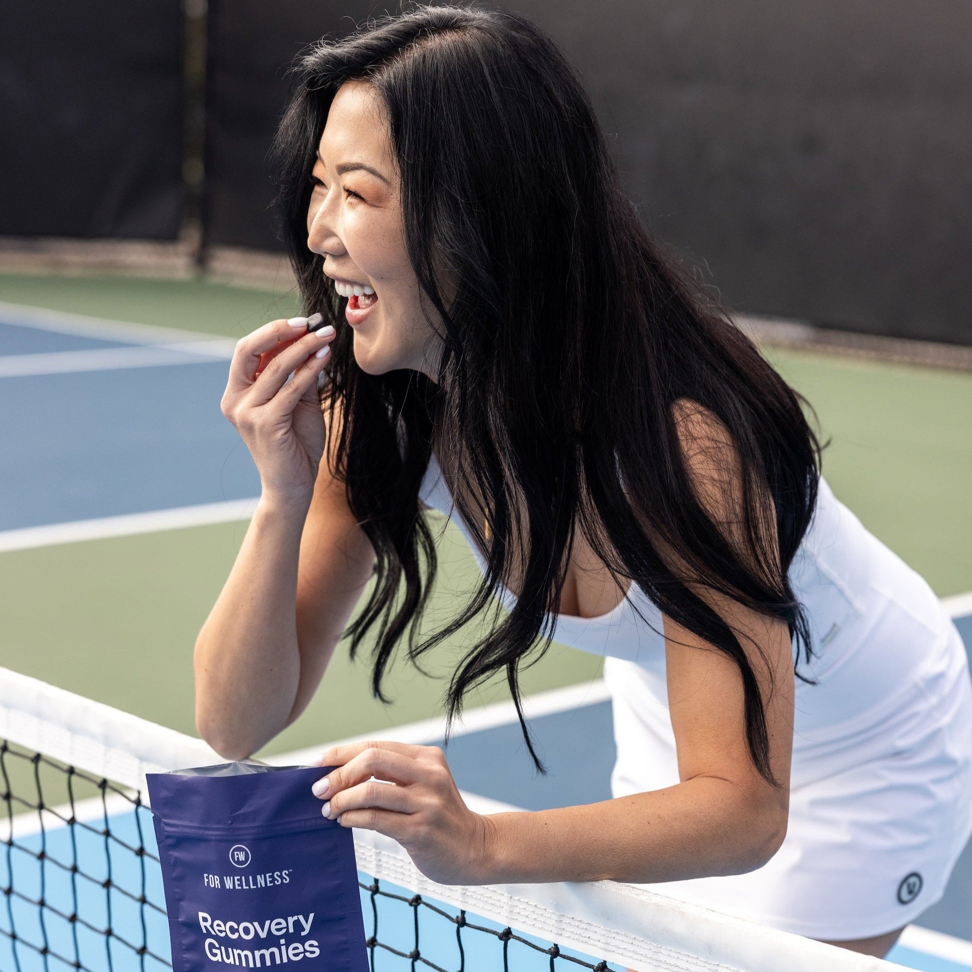 Smiling woman on a tennis court holding recovery gummies by the net.