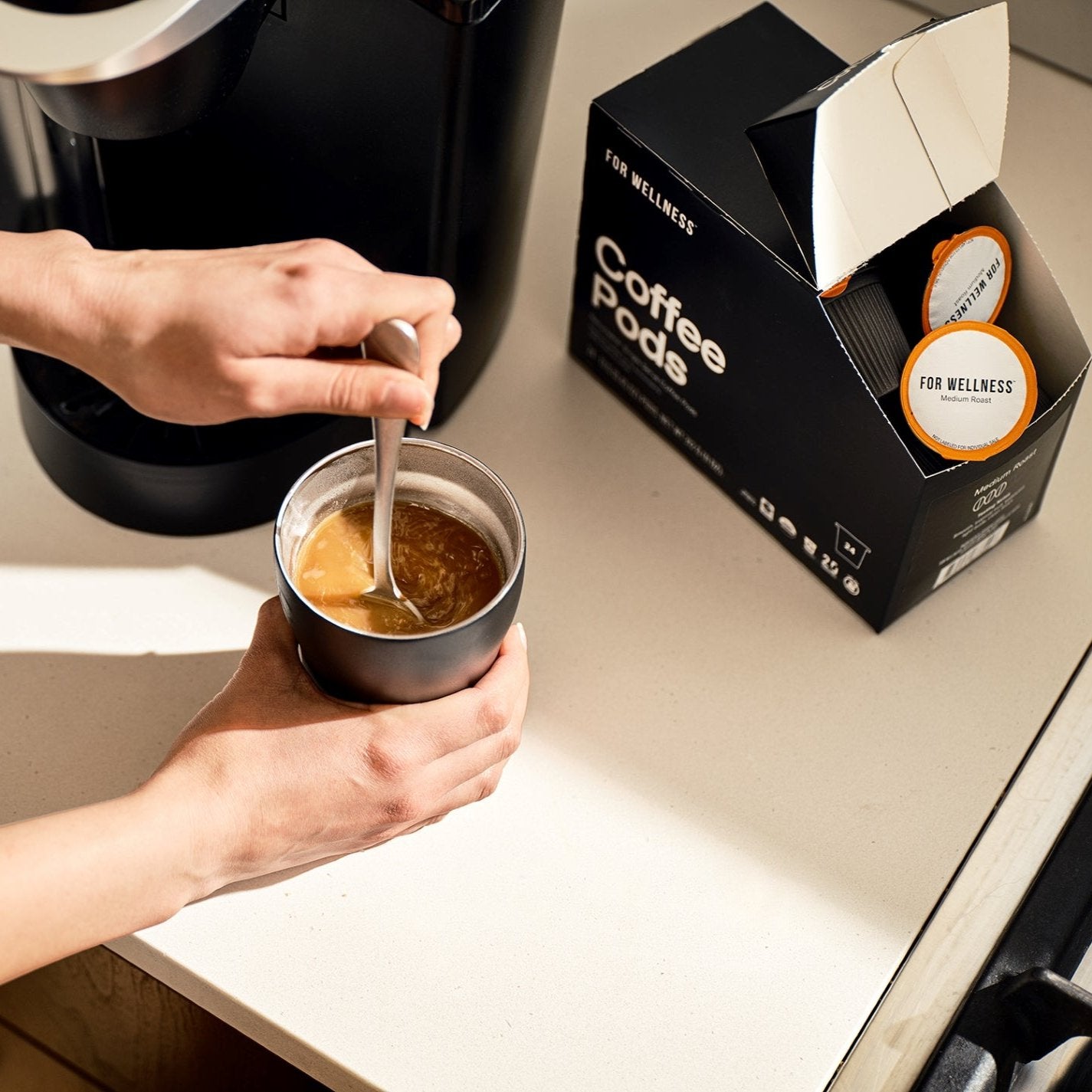 Person stirring coffee in a mug next to a box of coffee pods.