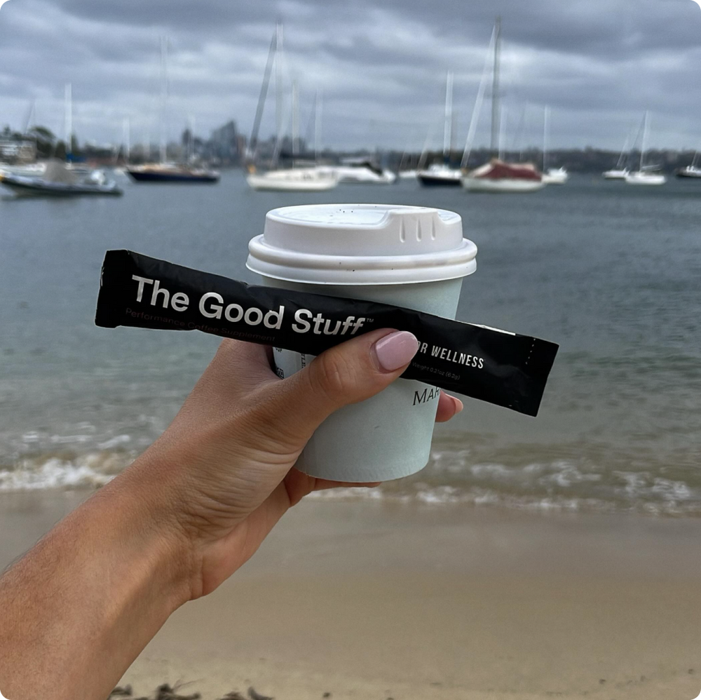 A hand holding up a cup of coffee and a travel pack of The Good Stuff on the beach 