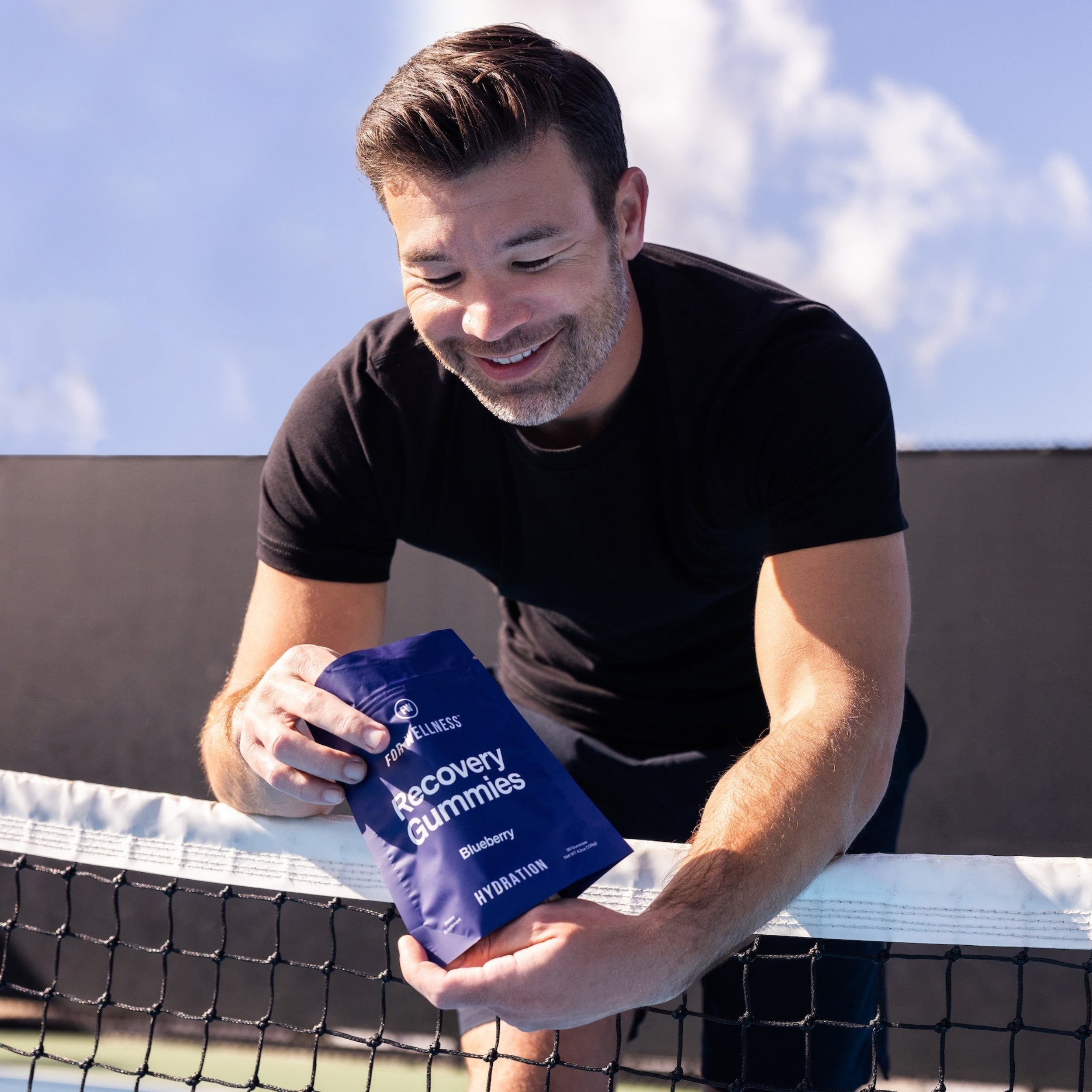 Man leaning over tennis net holding Recovery Gummies Hydration 